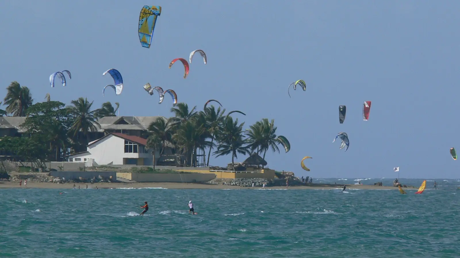 Una docena de cometas de kitesurf sobre la bahía de Cabarete, con riders en el agua y casas bajas entre palmeras en la punta.